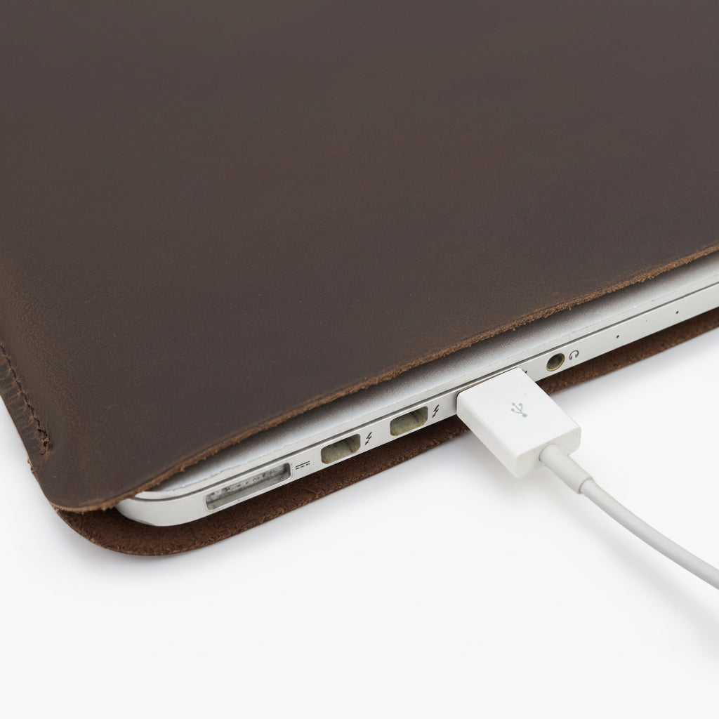 MacBook inside a dark brown leather sleeve with charging cable connected and ports accessible.